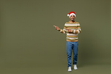 Fototapeta na wymiar Full body young african man in knitted sweater red Christmas Santa Claus hat point finger aside on area mock up isolated on plain green khaki background studio Happy New Year 2022 celebration concept.
