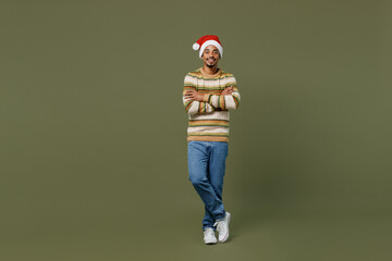Full body young african man in knitted sweater red Christmas Santa Claus hat hold hands crossed folded isolated on plain green khaki background studio portrait Happy New Year 2022 celebration concept