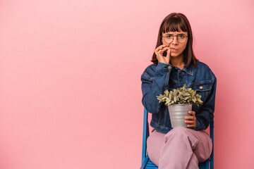 Young mixed race woman sitting on a chair holding a plant isolated on pink background with fingers on lips keeping a secret.