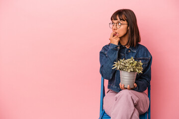 Young mixed race woman sitting on a chair holding a plant isolated on pink background relaxed thinking about something looking at a copy space.