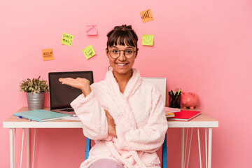 Young mixed race woman preparing a exam in the room wearing pajama isolated on pink background showing a copy space on a palm and holding another hand on waist.