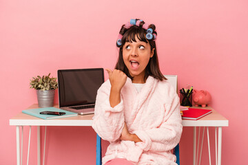 Young mixed race woman in pijama and with curlers isolated on pink background  points with thumb finger away, laughing and carefree.