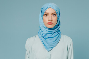 Young serious calm confident arabian asian muslim woman in abaya hijab clothes look camera isolated on plain blue color background studio portrait. People uae middle eastern islam religious concept.