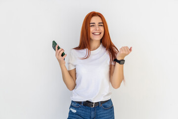 Fototapeta premium Portrait of redhead woman celebrating, shouting yes with satisfaction, fist pump, holding smartphone, winning on mobile phone, achieve daily goal in application over white background