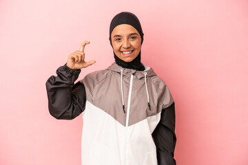 Young Arab woman with sport burqa isolated on pink background holding something little with...