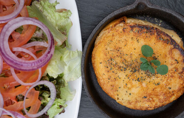 Top view of grilled provolone cheese served in a black pan and a fresh salad with tomato, red onion...