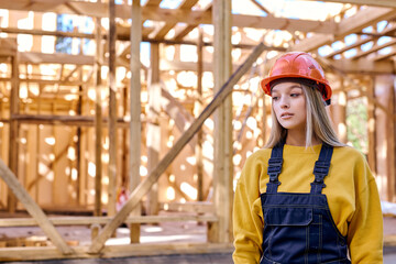 young caucasian builder architect female in hardhat posing against New residential construction home framing, at sunny day outdoors, dressed in working uniform and orange helmet, look at side