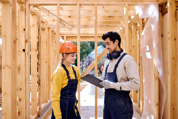 Fototapeta na wymiar Architect and carpenter discussing the construction plans while standing in unfinished house, female architect and male builder dressed in working uniform have talk, share opinion, building cottage