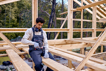 Young Hardworking Caucasian Contractor Working With Drill Driver. Carpenter Working on Wooden House Skeleton Frame Roof Section. Construction Industry Theme. Bearded Male Is Focused On Work