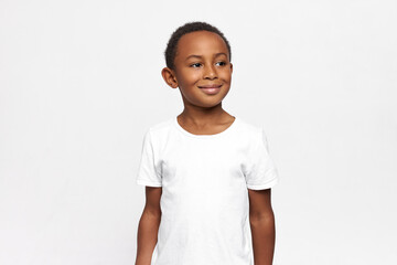 Good-looking dark-skinned elementary school boy wearing white T-shirt with copy space for your advertising content, smiling looking aside. Carefree childhood. Happy as kid. Human emotions and feelings © Anatoliy Karlyuk