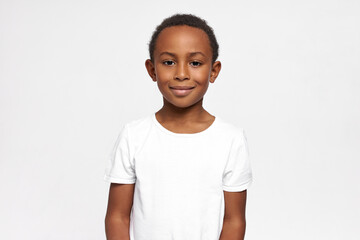 Happy kid, looking at camera with content smile, feeling proud of himself and his school marks, dressed in white casual T-shirt, having curly short haircut, posing with delight face expression