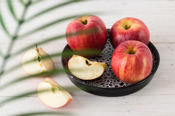 In a round black dish on white wooden table are three juicy beautiful ripe red apples of the early winter variety Gala. Next to the slices of the cut fruit. Top view through a palm leaf