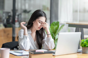 Obraz premium Young Asian woman feeling migraine head strain. Tired, Overworked businesswoman financier while working on laptop computer in office.