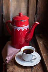 A white cup with black coffee is staning near a red coffee pot on a wooden window sill by the window. Breakfast theme.