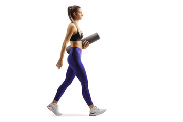 Full length profile shot of a young female in crop top and leggings walking with an exercise mat - obrazy, fototapety, plakaty