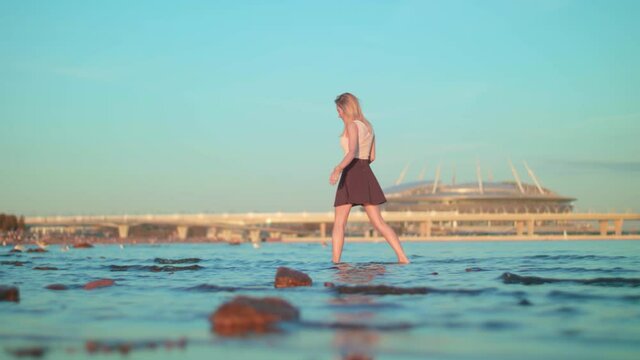 European sunburnt beautiful attractive blonde skinny fit girl in brown skirt and white t-shirt walks on the water near the shore with stones at evening sunset with blue sky background Gazprom Arena