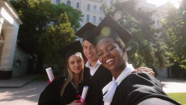 Group of multiracial students graduates two ladies and one guy taking selfie video with the camera smiling large after the graduation