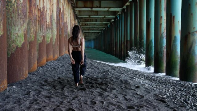 a barefoot woman dances under the pillars of the bridge against the background of the incoming waves. she has turned back and is trampling. the camera is moving. the general plan