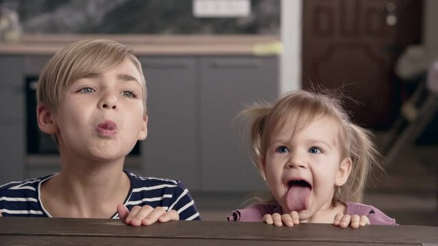 Children look out from the table, laugh and stick out their tongues, slow motion.