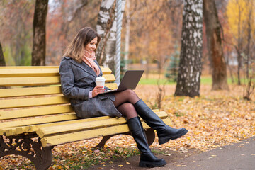 Girl sits on a park bench in autumn with a laptop and drinks coffee