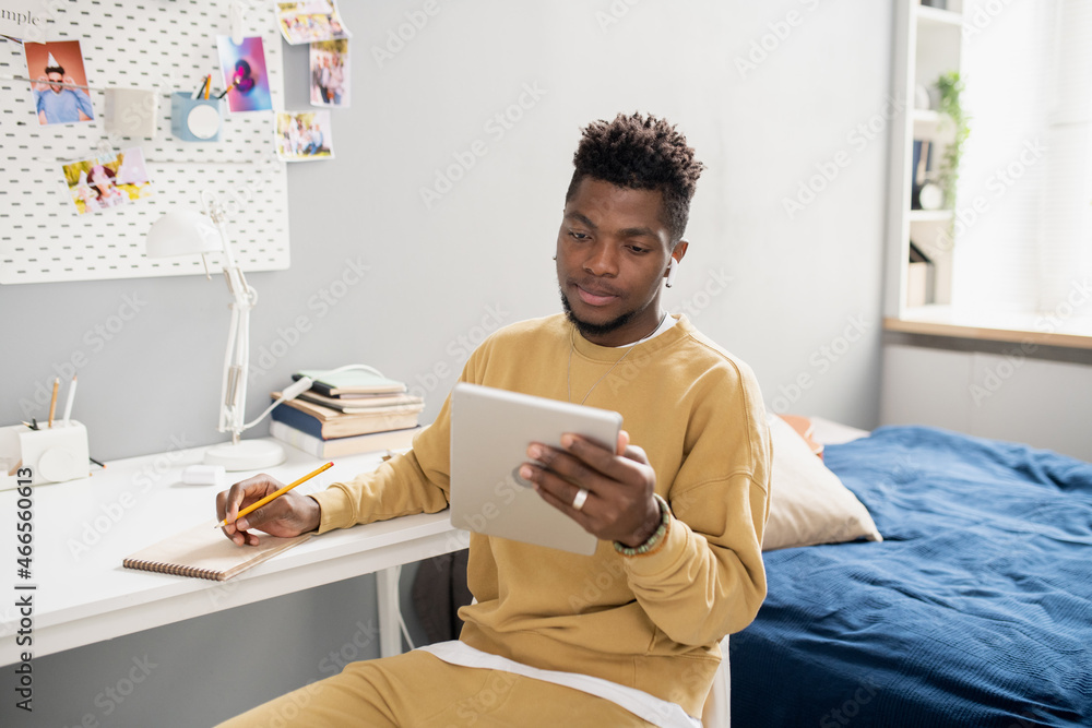 Poster young serious student in casualwear looking at tablet screen and making notes in notepad during onli