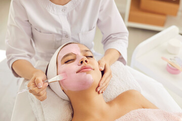 Happy calm relaxed beautiful young woman or teenage girl lying on soft towel in beauty parlor or spa salon and enjoying soothing pampering pink kaolin clay facial mask for fresh clear skin, close up