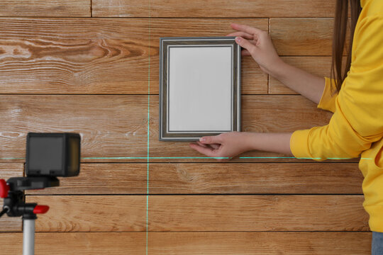 Woman using cross line laser level for hanging blank photo frame on wooden wall, closeup