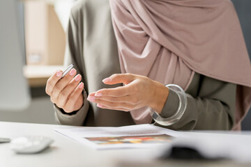 Hands of young teacher in hijab sitting by desk and explaining subject during online lesson or training
