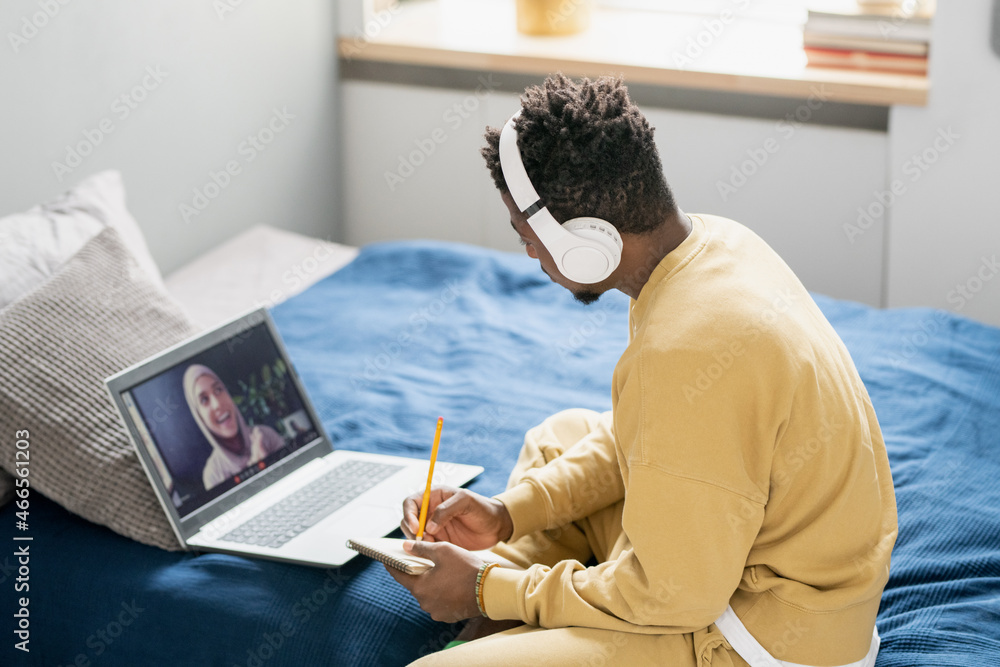 Poster contemporary student of african ethnicity sitting on bed and making notes in notepad while looking a