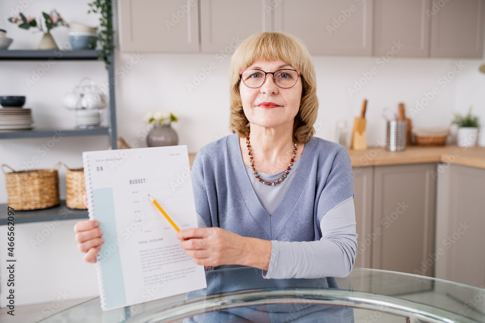 Poster confident blond female pointing at financial paper while explaining subject by kitchen table in fron