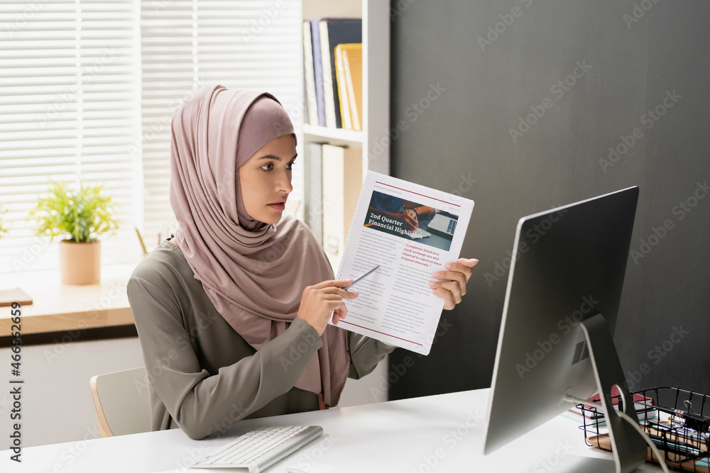 Poster confident teacher of economics pointing at financial document while sitting in front of computer mon