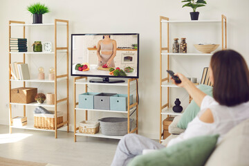 Young woman watching television sitting on sofa at home. Adult lady with remote switches vegan or vegetarian cooking TV show on with girl making lunch or dinner, cutting veggies and preparing salad