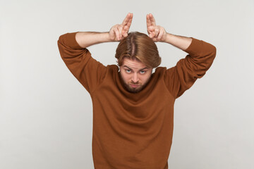 Portrait of angry bully man with beard wearing sweatshirt, showing bull horn gesture with fingers...