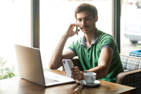 Portrait Of Busy Handsome Businessman Wearing Green T-shirt, Working On Laptop, Talking On Mobile Phone And Looking At Another Cellphone. Indoor Shot Near Big Window, Cafe Background.