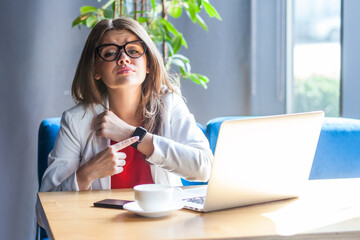Portrait of unhappy woman in glasses pointing to smartwatch on her hand, looking gloomy...