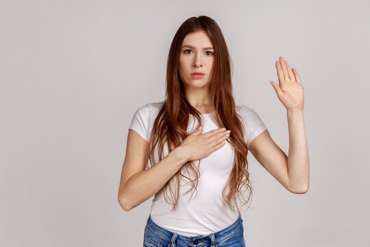 Portrait of very responsible and honest woman giving promise, making solemn vow in ceremonial tradition with raised hand, wearing white T-shirt. Indoor studio shot isolated on gray background.