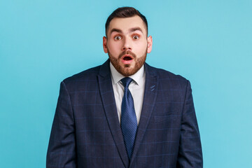 Wondered business man wearing official style suit looking at camera with shocked face and big eyes and open mouth, hearing astonishing news. Indoor studio shot isolated on blue background.