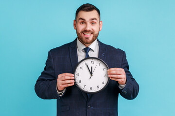Portrait of positive cheerful businessman wearing dark suit holding big clock and looking at camera...
