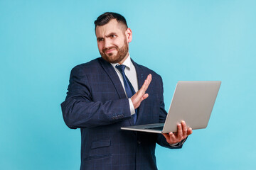 Portrait of attractive bearded young man wearing official style suit stretching hand, dont want to see, looking away, disgusting content, Indoor studio shot isolated on blue background.
