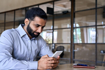 Smiling indian ceo businessman holding using cell phone mobile apps sitting at office desk looking...