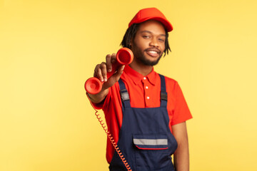 Portrait of happy positive handyman wearing blue uniform holding out handset, asking call him and order service, looking friendly at camera. Indoor studio shot isolated on yellow background.