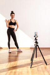 Slim female recording tutorial video for fitness vlog, waving hand, saying hi to followers, wearing black sports top and tights. Full length studio shot illuminated by sunlight from window.