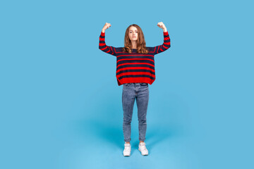 Full length of confident woman, standing with strong gesture and looking at camera, raised arms, wearing striped casual style sweater. Indoor studio shot isolated on blue background.