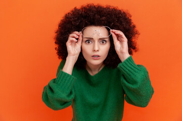 Surprised woman with Afro hairstyle wearing green casual style sweater standing raised her eyeglasses, looking attentively, sees something shocking. Indoor studio shot isolated on orange background.