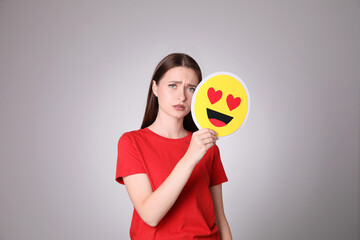 Sad young woman covering face with heart eyes emoji on grey background