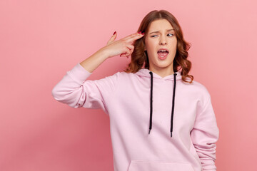 Portrait of depressed curly haired teen girl in hoodie pointing finger gun to head and looking desperate, making suicide gesture, shooting herself. Indoor studio shot isolated on pink background