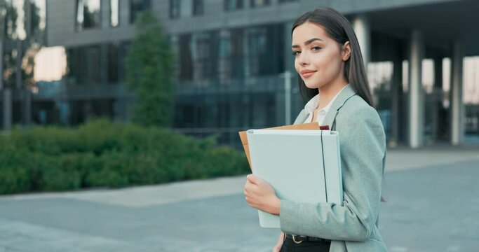 A smiling, pretty, successful girl running her own company manager boss is standing next to glass modern building going to meeting conference to work holding files with documents in her hand