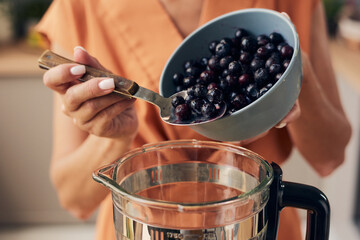 Hands of young woman putting fresh blueberries into electric blender while cooking homemade...