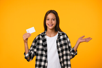 Happy smiling caucasian teenager schoolkid girl pupil student holding credit card for e-banking, online shopping, paying bills isolated in yellow background.