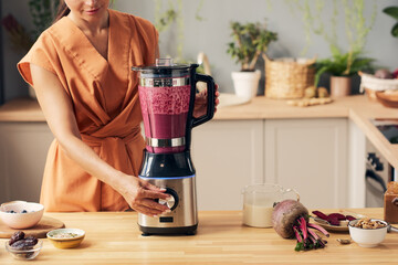Young housewife with electric blender preparing beetroot smoothie by kitchen table with bowls...